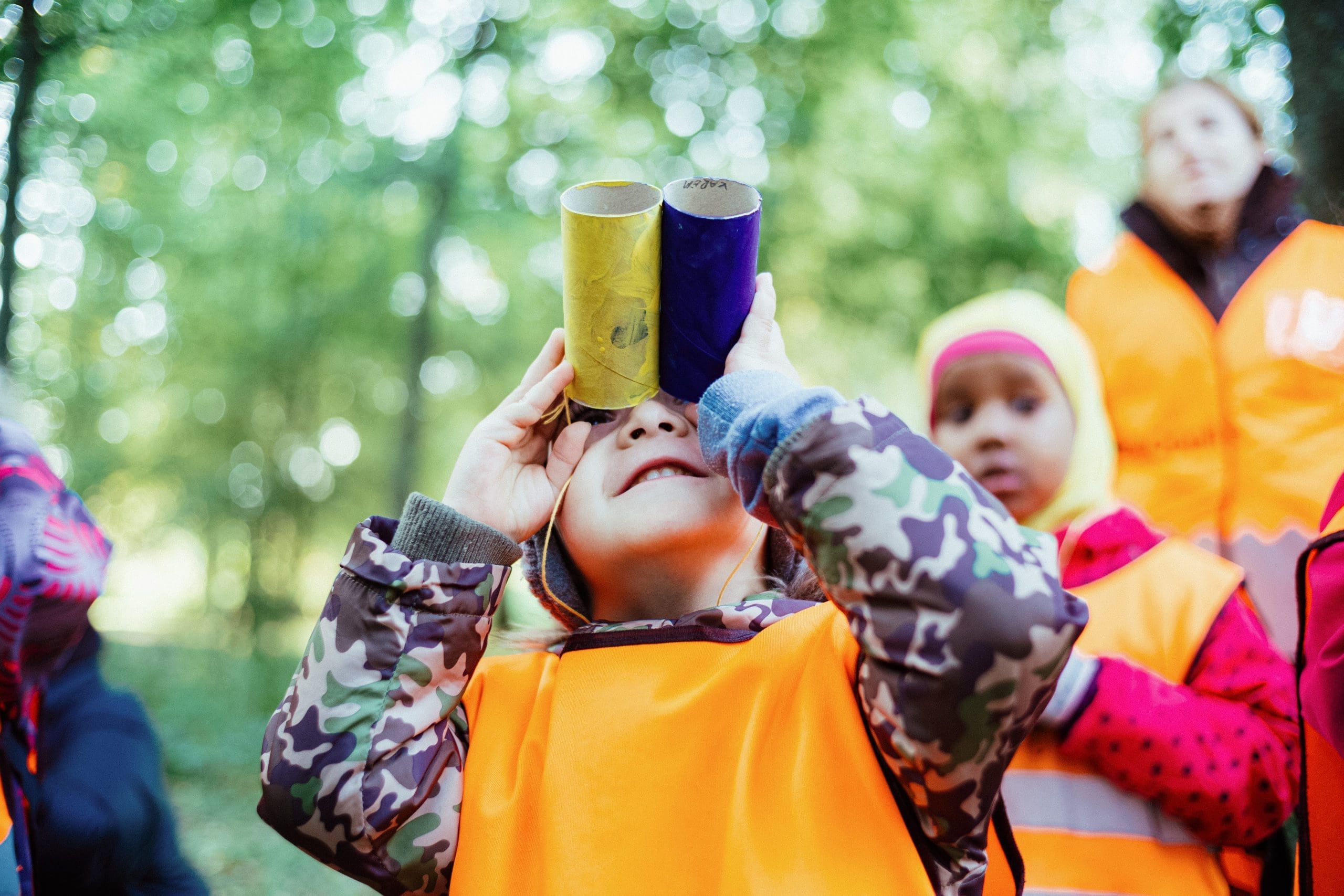 A kid facing the camera on a trip to the woods with daycare, looks up towards the trees through homemade binoculars whilst another kid and teacher watch in the background.