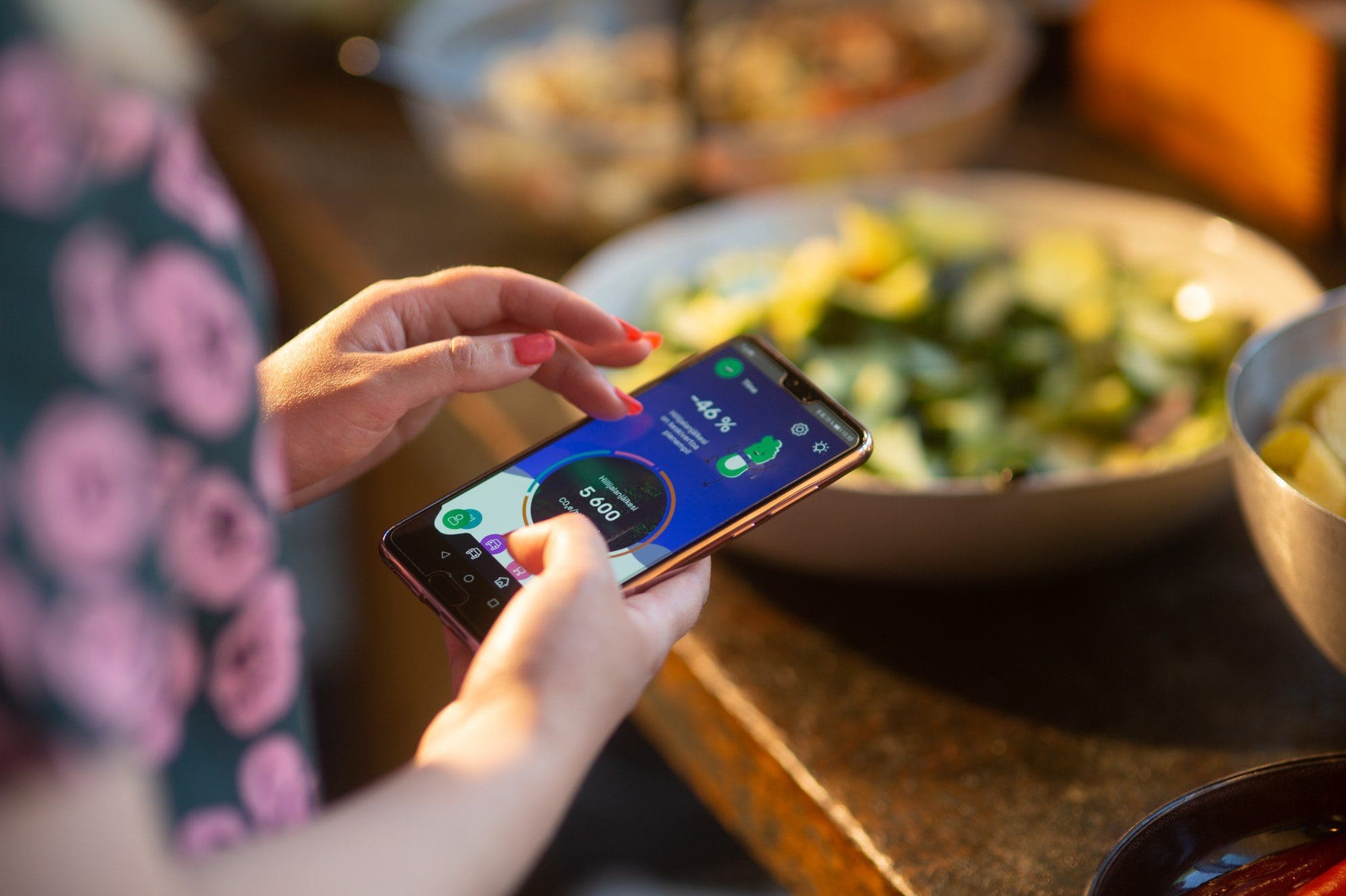 Viewed from behind and over their right shoulder, a woman is using the Carbon Ego app on her phone, standing in front of a buffet table.