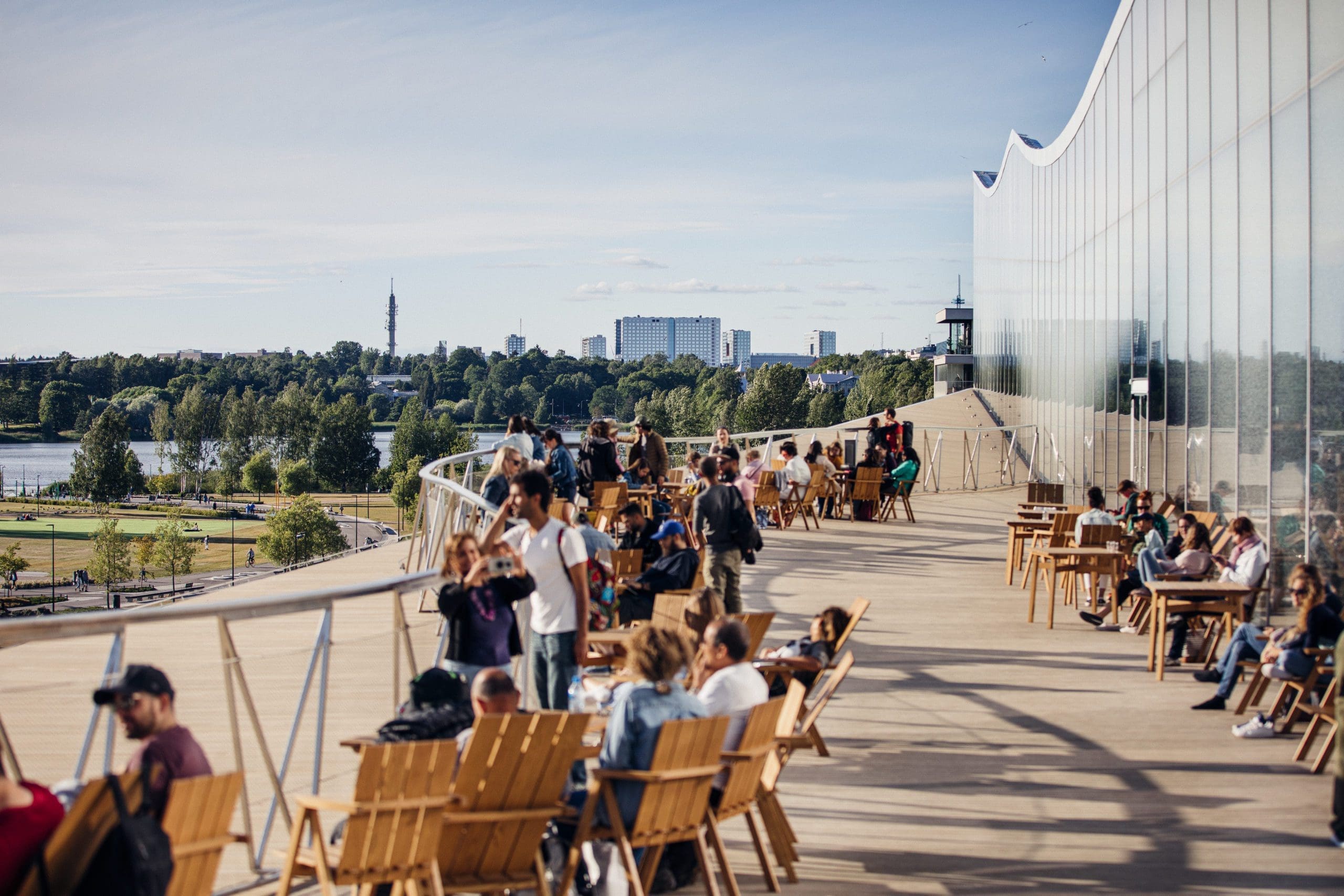 A small crowd of people sit in deck chairs and stand by the railings, enjoying the sunny weather whilst admiring the view from Oodi's summer terrace.