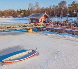 A view from the the frozen water looking towards land and Cafe Regatta in winter. Everything is covered by a light dusting of snow, a boat is sitting in the ice in the bottom left of the photo, and the trees of Sibelius Park cover the horizon in the background. People are queuing to get into the cafe.