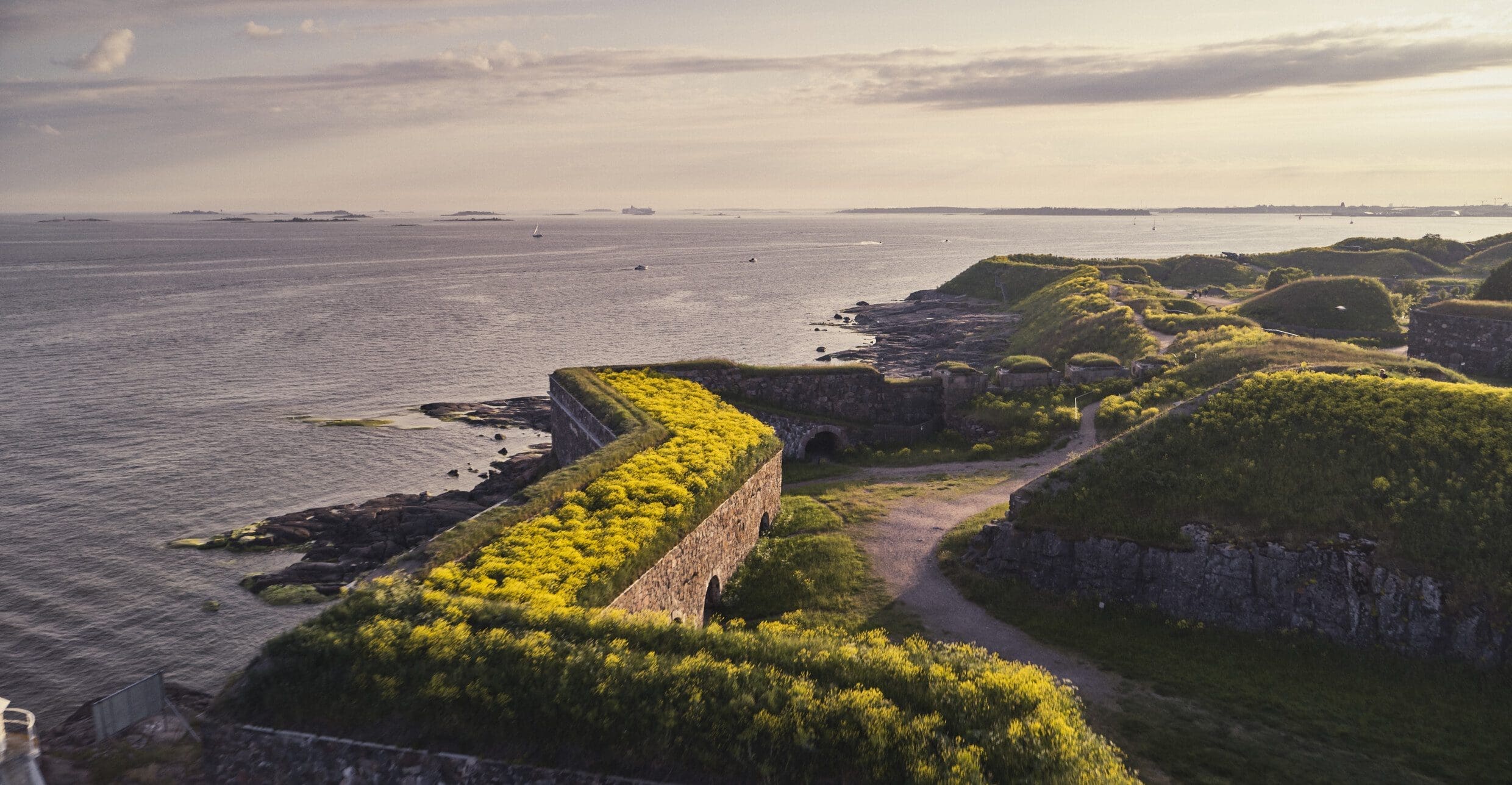 A view from above looking down on some of the star points of Suomenlinna's remaining fortress walls. The island is very green and in full bloom, the sea stretching to the horizon under a sunny blue sky.