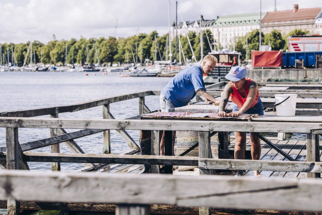 Two people are scrubbing rugs at the mattolaituri on the edge of Kaivopuisto, the boats and shoreline of Merisatama in the distance.