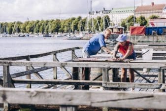 Two people are scrubbing rugs at the mattolaituri on the edge of Kaivopuisto, the boats and shoreline of Merisatama in the distance.