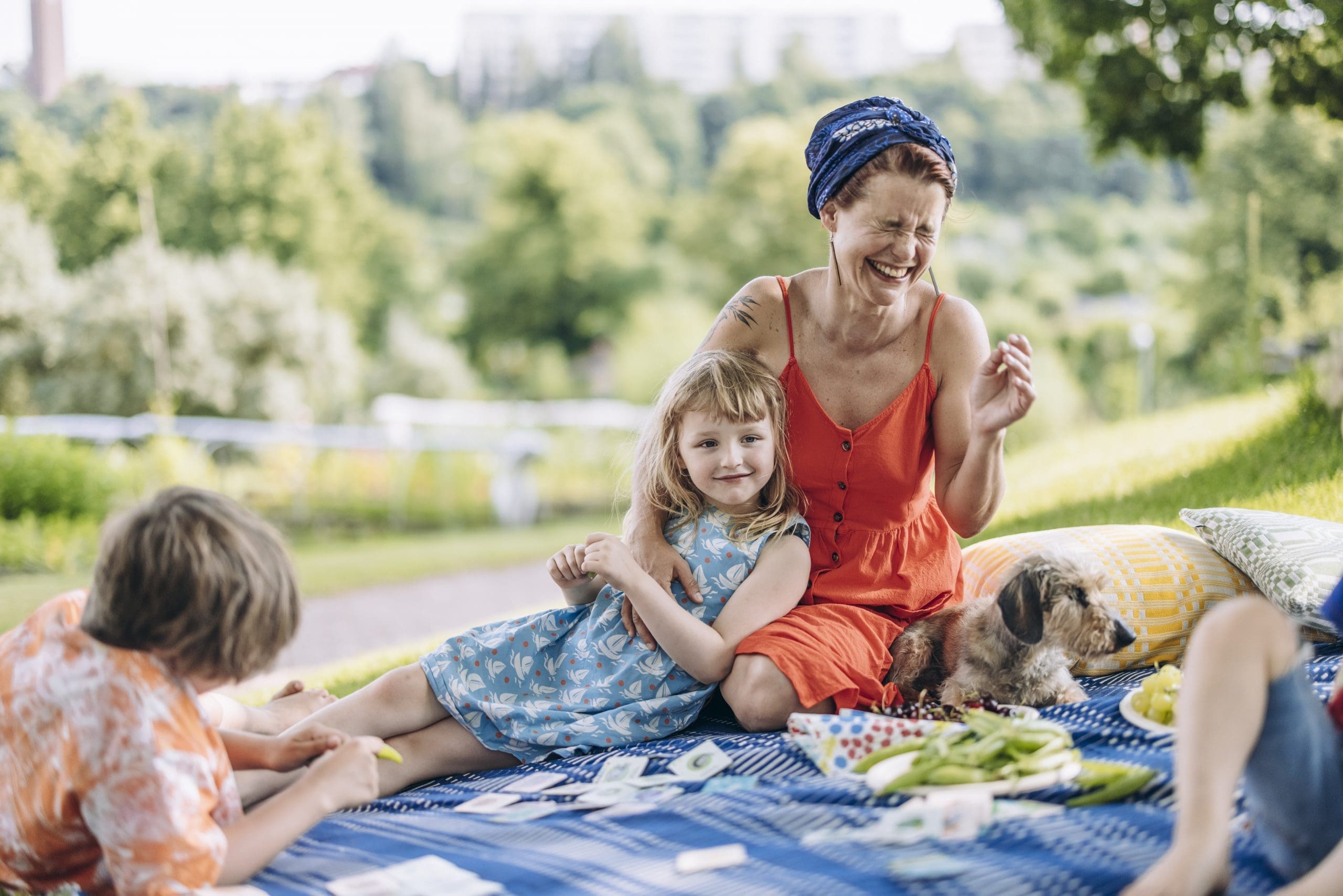 Sitting on a picnic blanket with the rolling greenery of the park in the background, a mother laughs with their eyes closed while holding a girl who's lying in her lap. Two other kids and a small dog are also laying on the blanket.