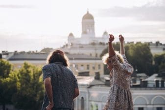 With their backs facing the camera and standing on a hill in front of Uspenski Cathedral, the man on the left faces forward while the woman on the right reaches her arms into the air whilst smiling. In the background, rooftops and trees can be seen, Helsinki Cathedral sitting on the horizon.