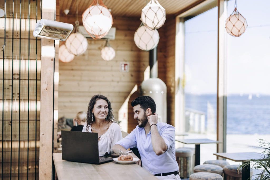 A man and woman sit discussing in front of a laptop at the end of a long table in cafe Birgitta. The walls are natural wood, spherical lamps wrapped in fishing nets hang from the ceiling and to the right are floor to ceiling windows showing the sea underneath a blue sky.