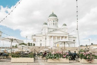 The Senate Square has been transformed into the summer terrace, which is full of seating under parasols as well as wooden boxes bursting with pink and white flowers. The Helsinki Cathedral is sitting in the background against a cloudy blue sky.