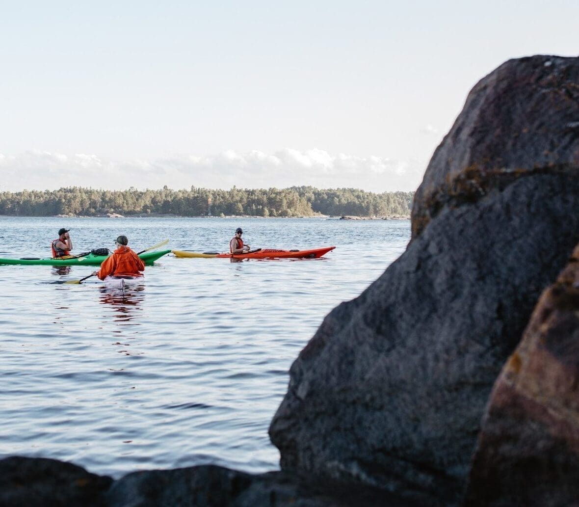 In the foreground to the right, some rocks partially obscure the view of four people canoeing to the left, the tree covered shoreline can be in the distance under a blue sky.