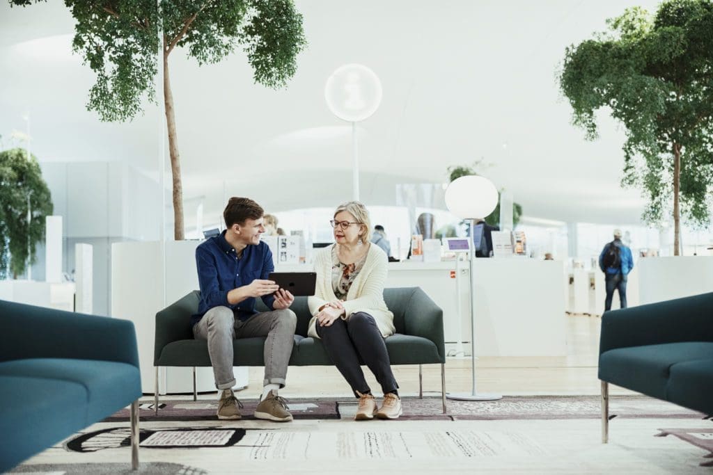 With an Oodi service desk behind the sofa they're sitting on, a man uses a tablet whilst offering customer service to an older woman.