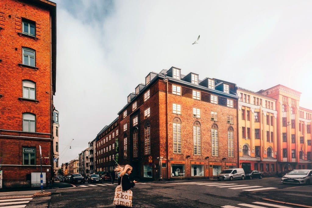 View of Hotell Anna from across the street, cars are using the crossroads driving past the red brick buildings of the area. A woman is crossing the street in the foreground.