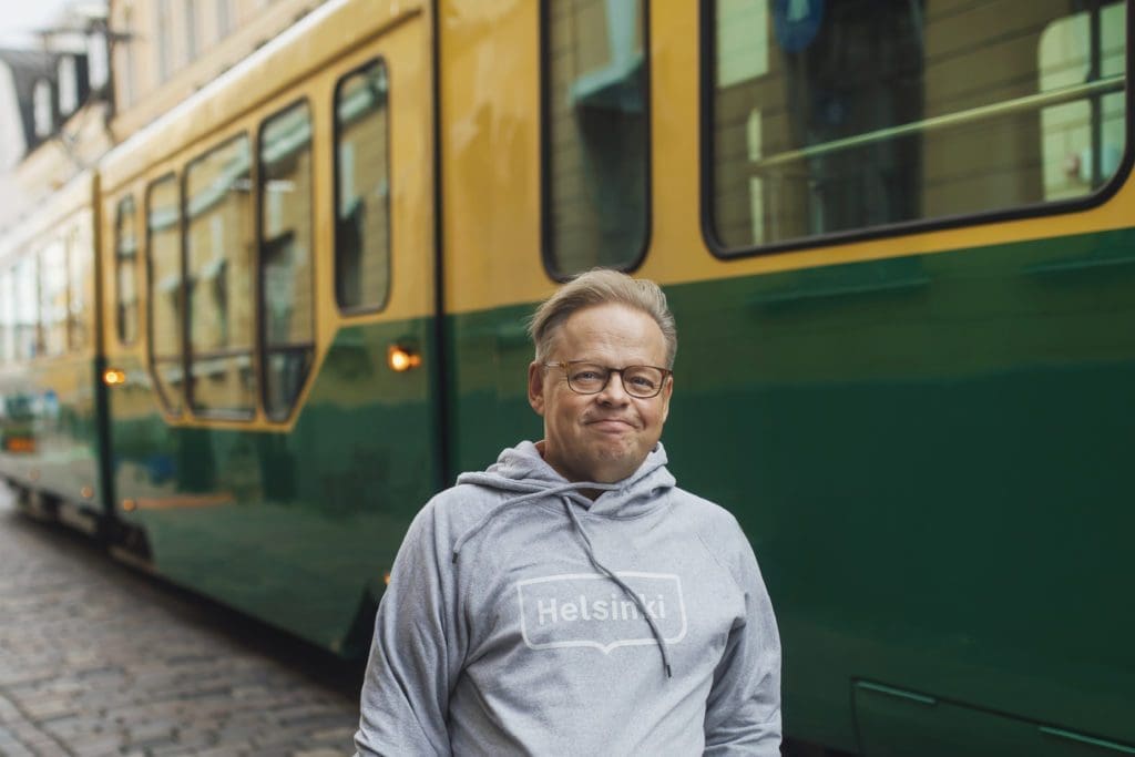 Helsinki Mayor, Juhana Vartiainen, wearing a Helsinki hoodie, looks towards the came and smiles a tram passes by behind him.