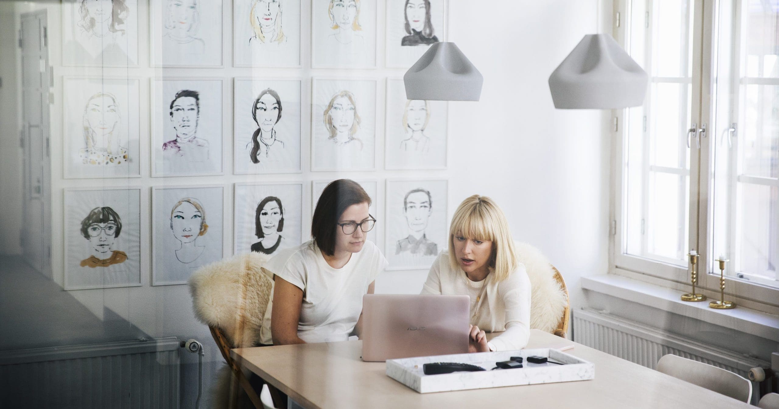 Viewed from behind an office window which shows slight reflections, two women are sitting at a table discussing something that is on a laptop in front of them. The office wall behind them is covered in stylised, illustrated portraits.