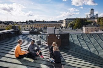 It's a sunny day where two men and two women are sitting casually on a Helsinki rooftop having a meeting. The tops of other buildings can be seen in the background with Helsinki Cathedral in the distance on the right.