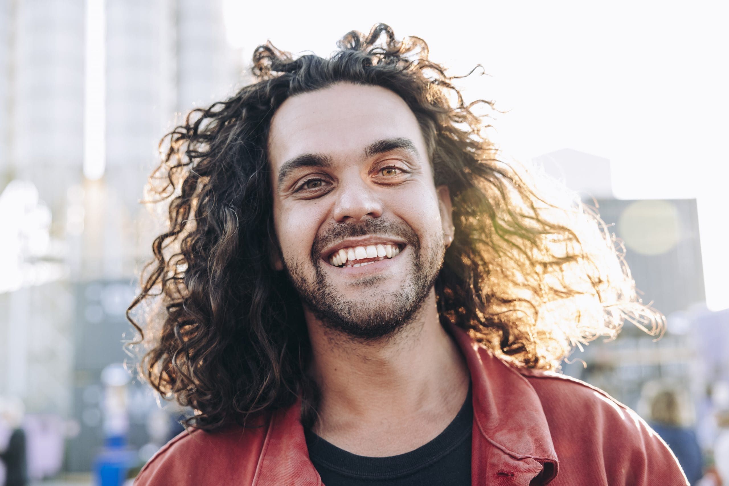 Against a busy and blurred background, a very smiley, slightly bearded man with long, brown hair and a red jacket is looking straight at the camera. Only the head and shoulders are shown.