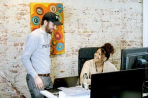 A woman sitting at her desk, is smiling with a male colleague to the left. The wall behind them is roughly finished red brick and holds a colourful abstract painting.