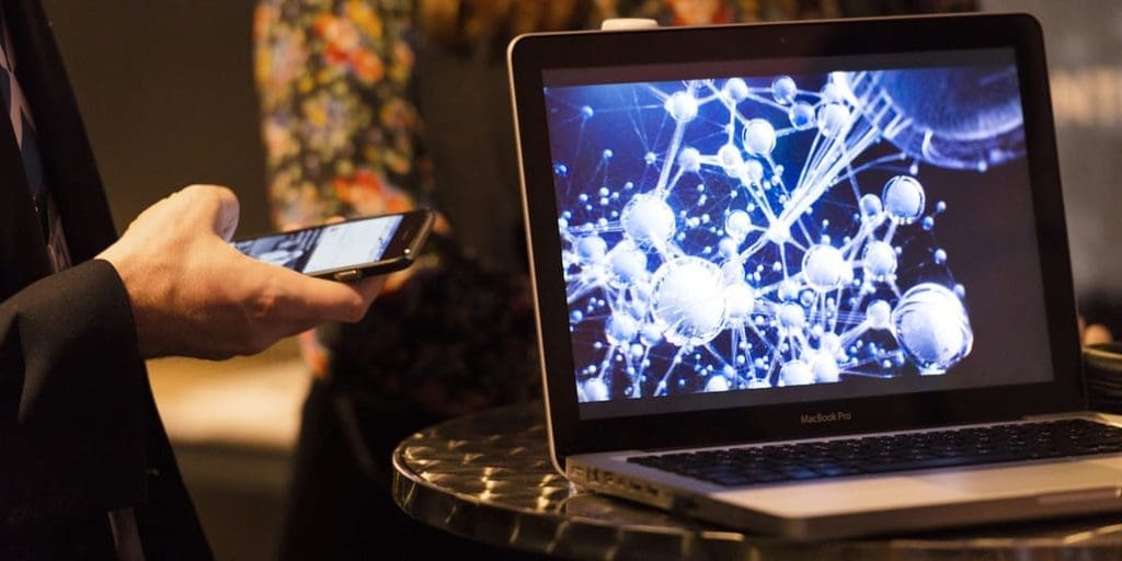 Close up of a laptop sitting on a standing table to the right, on the is left someone's hand holding a phone whilst they are using it.