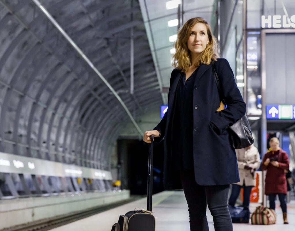 Standing in the huge underground tunnel that is the Helsinki airport tain station, a woman wearing a long blue coat stands at the platform waiting for her train into Helsinki. On the left are the train tracks and on the right are the lifts and escalators to the airport.