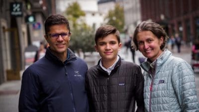 Carr family and 90 Day Finn participants- Sean, Sebastian and Ladi are standing and smiling at the camera with part of the Stockmann building and pedestrian areas of Helsinki in the background.