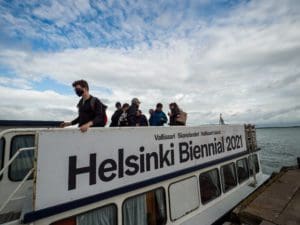 90 Day Finn participants are waiting to take their seats on top of a ferry to Helsinki Biennial 2021, held Vallisaari island. The boat is at dock with a partially cloudy and blue sky behind them.