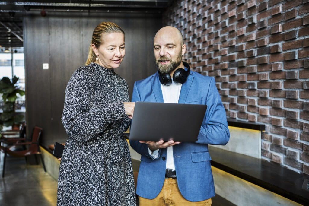 Standing inside an open office space with a red brick wall behind them, a man and a woman stand talking whilst looking and pointing at a laptop the man is holding.