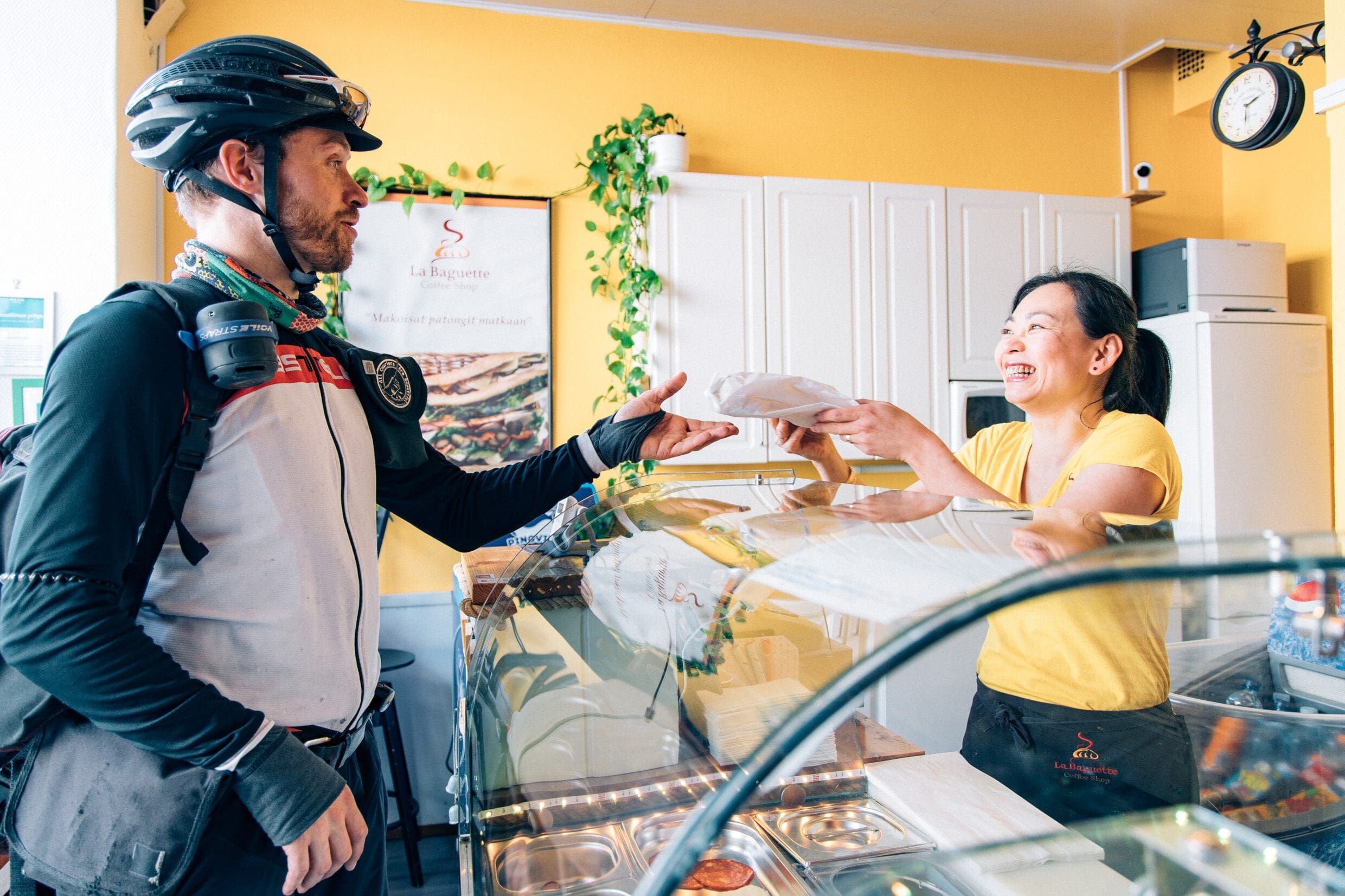 Inside a cafe with bright yellow walls, on the right a café owner passes a food order over a glass counter to a courier dressed in full cyclists' gear. The woman is smiling broadly.