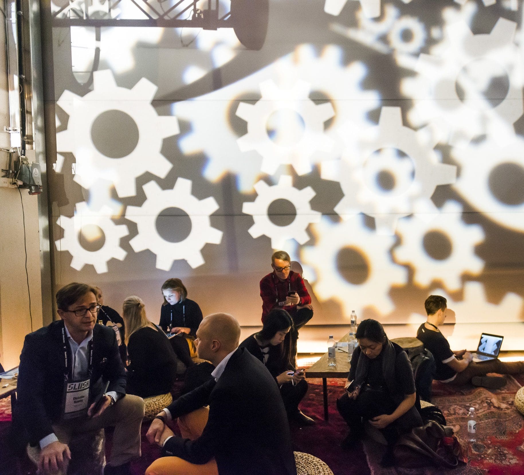 In an event at Slush, a group of business people are sat on large cushions on the floor as they discuss between themselves, while a large projection of cogs is being projected on the wall of the tall space behind them.