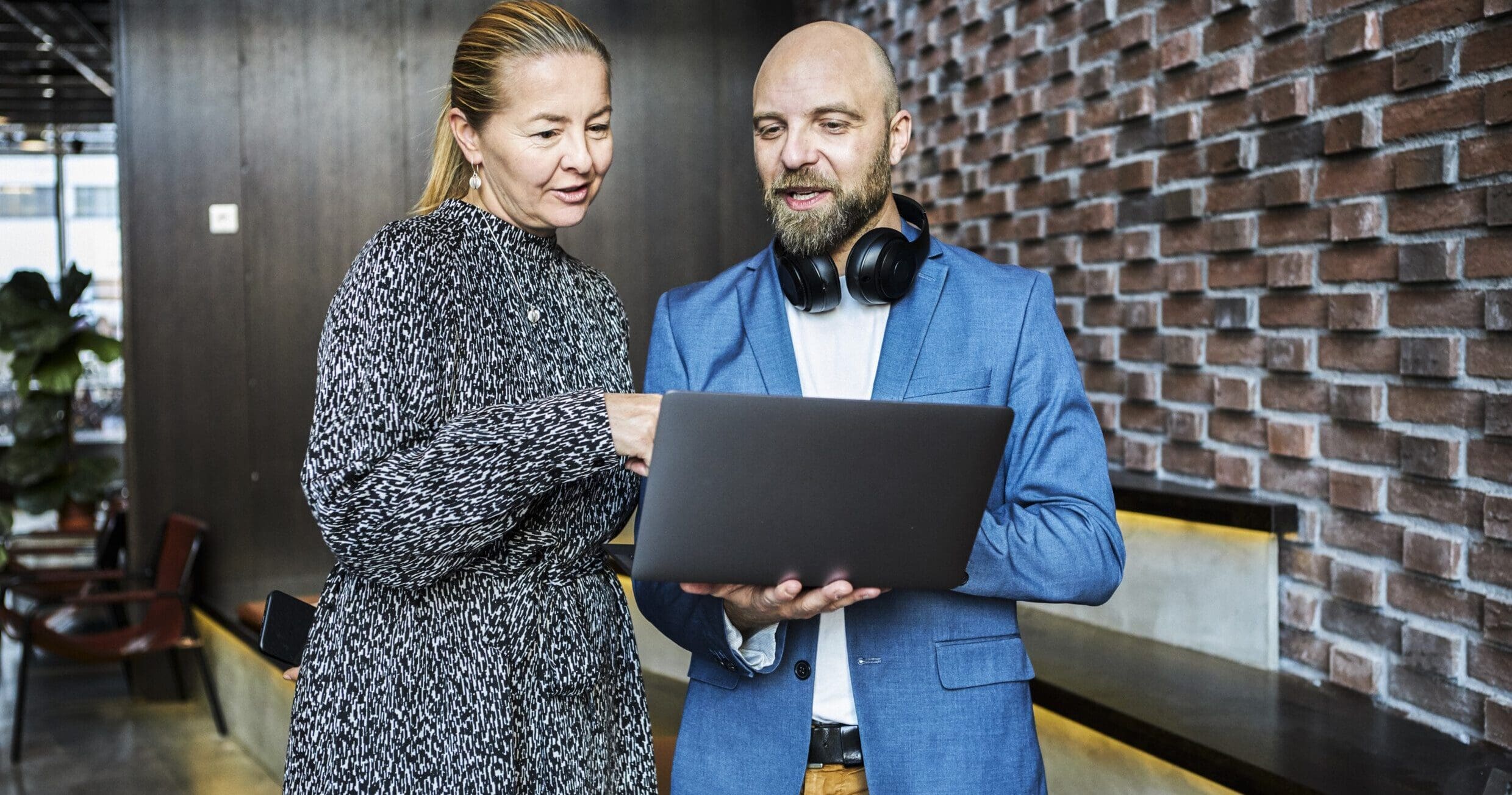 Standing inside an open office space with a red brick wall behind them, a man and a woman stand talking whilst looking and pointing at a laptop the man is holding.
