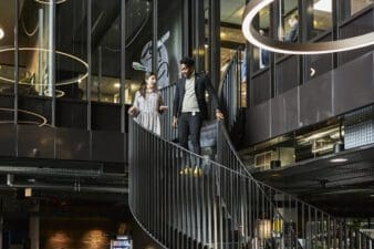 A man and a woman go down on spiral stairs talking with each other. The picture is taken in a modern office environment with rather black and silver tones.