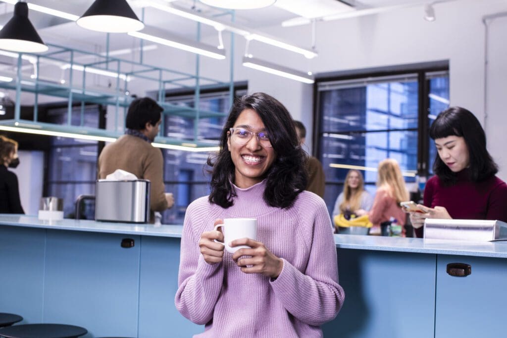 A person dressed in a pink sweater with black shoulder length hair, a big smile and a cup of coffee in her hands. She is in an office environment and in the background 3 other people are visible but not in the focus.