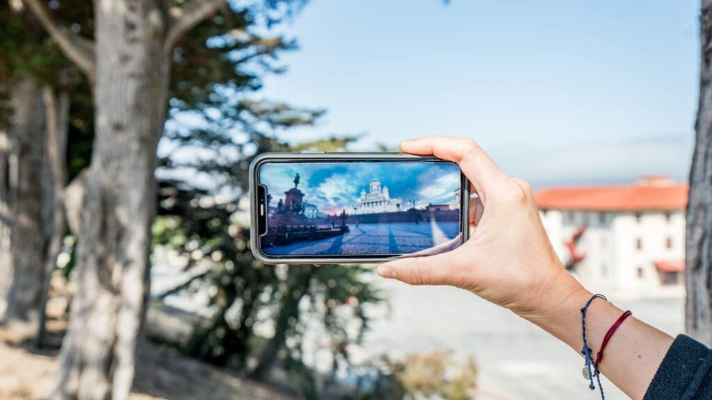 Against a sunny background with trees, blue sky and some buildings in the background, a hand holds a phone sideways as it shows a scene of the Senate Square and Helsinki Cathedral from Virtual Helsinki.