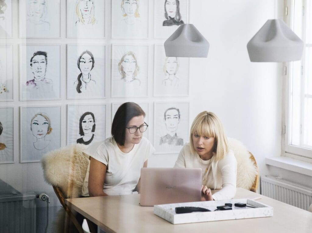 Viewed from behind an office window which shows slight reflections, two women are sitting at a table discussing something that is on a laptop in front of them. The office wall behind them is covered in stylised, illustrated portraits.