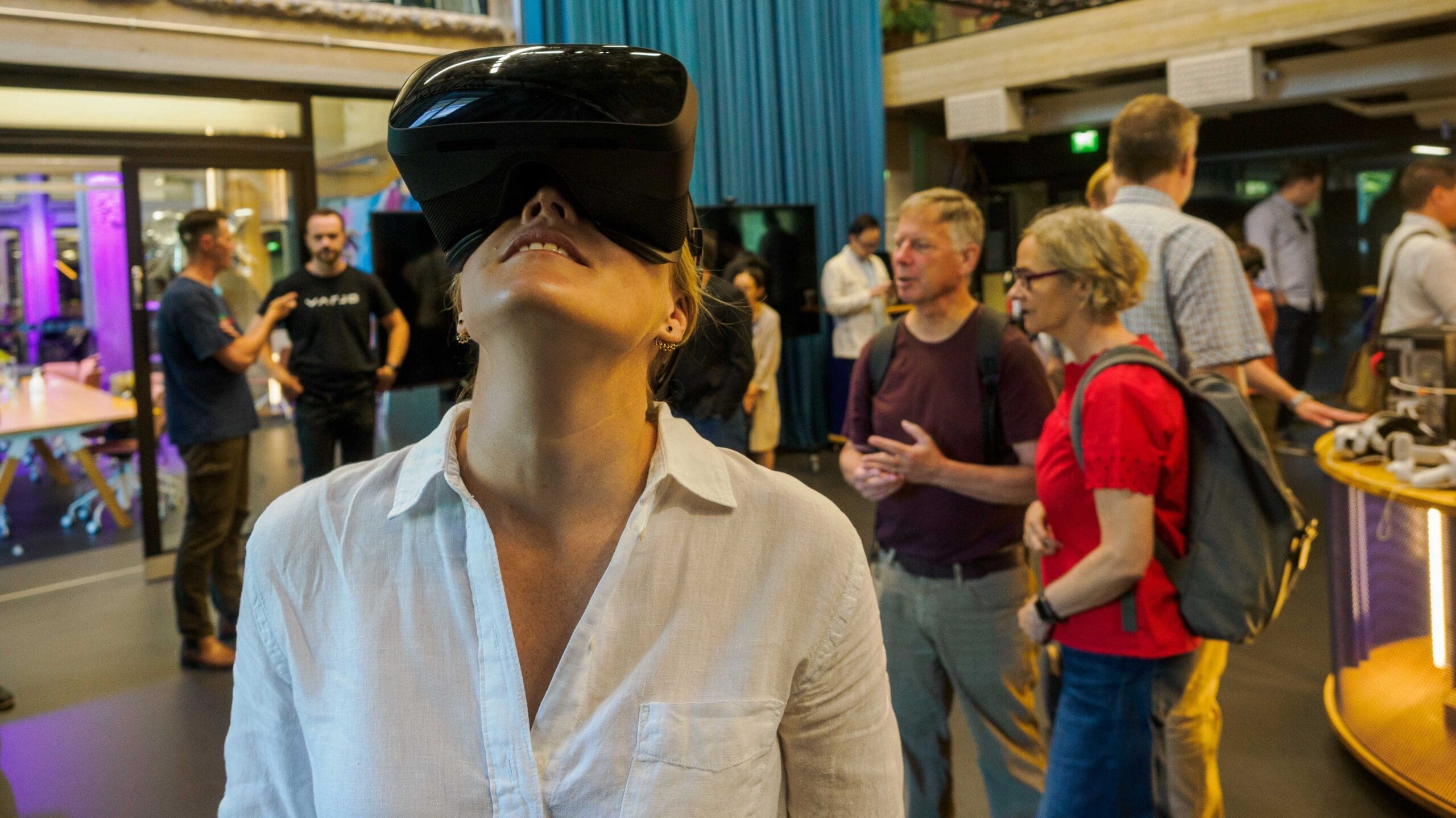 Woman looking up with the black VR glasses. She is wearing a white shirt and in the background one can see other people mingling.