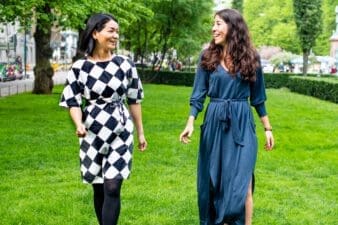 Two women looking at each other and smiling, outdoors, in the park. They are both dressed in summer long dresses. The park is all green and could be seen in the background.