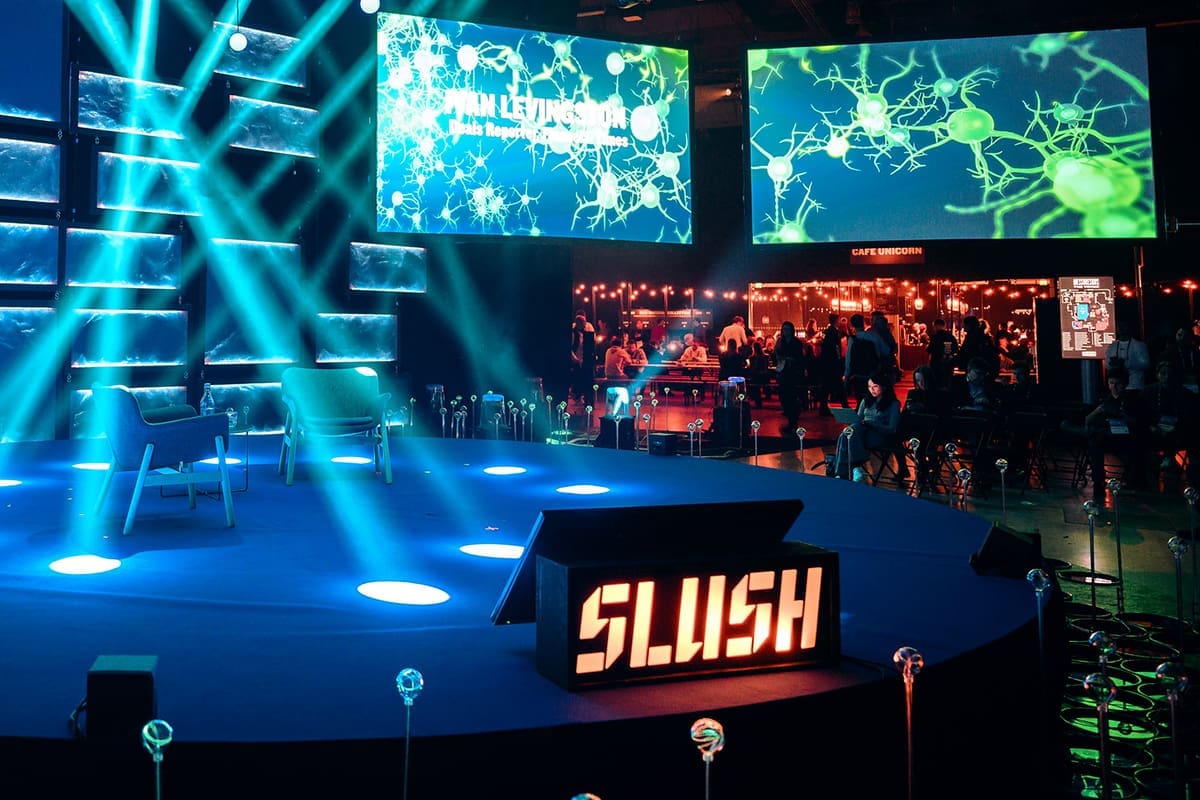 Slush sign placed on the front of the empty stage and on the left a crowd of people are networking.