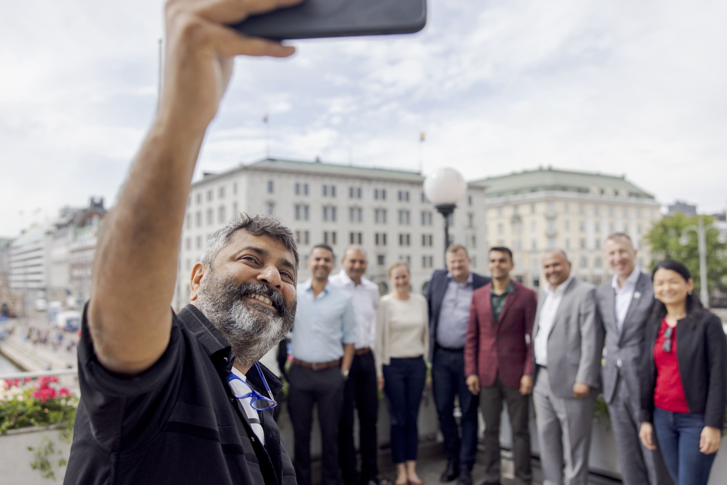 A man holds a phone above his head to snap a selfie of himself and a group of people behind him.